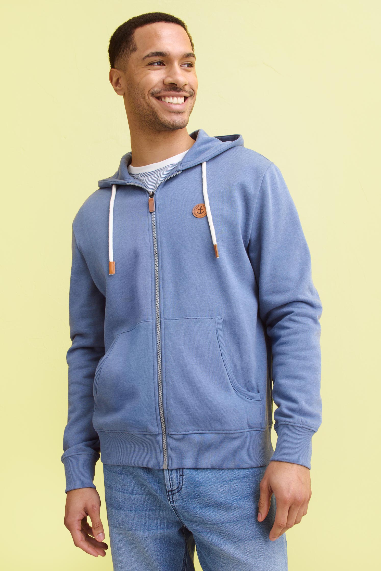 Hellblauer Kapuzenpullover mit Reißverschluss, weißen Kordeln und kleinem Logo. Dazu blaue Jeans.