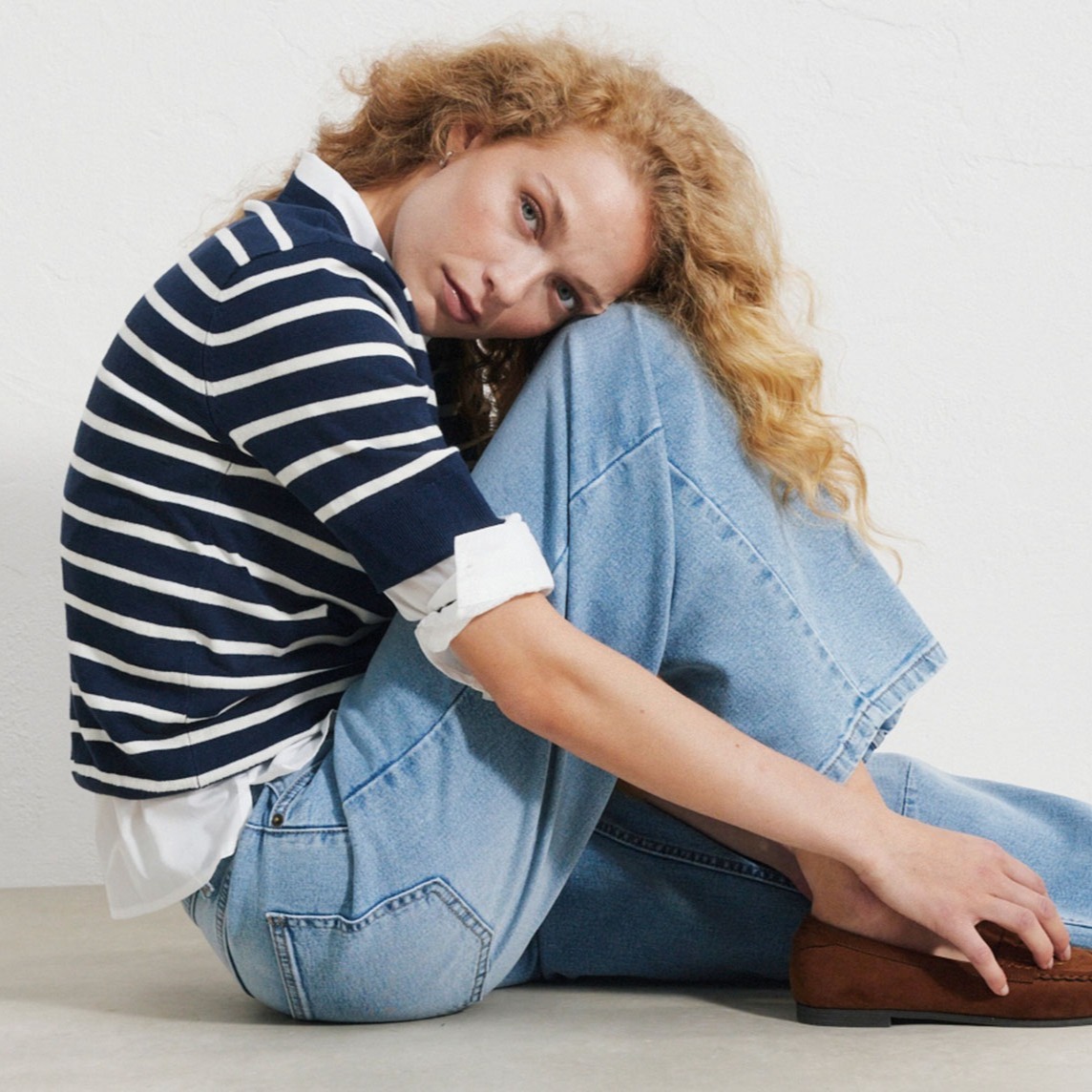 Gestreifter Pullover über weißem Hemd, weite blaue Jeans und braune Loafer.