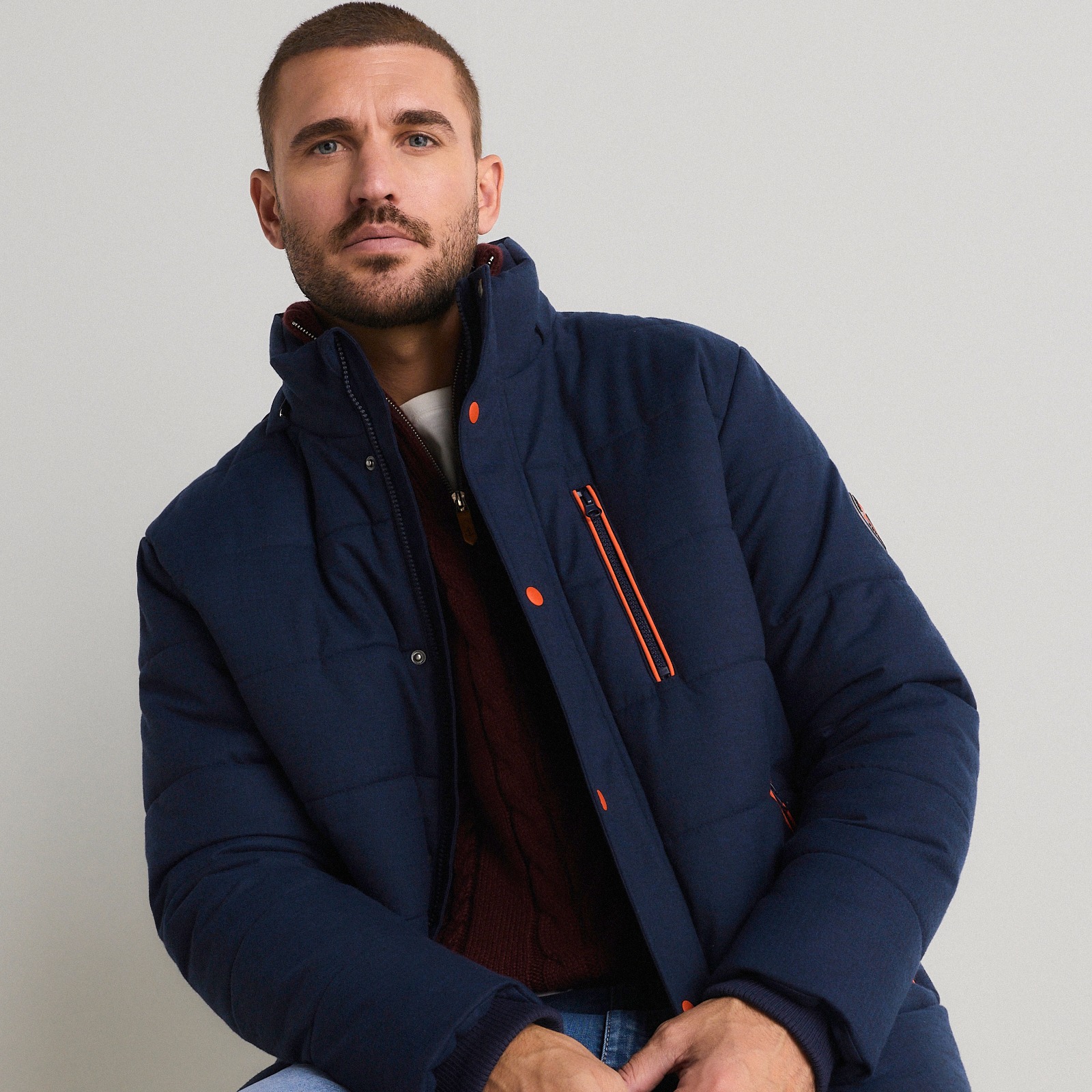 Der Mann trägt eine dunkelblaue Steppjacke, einen weinroten Pullover und hellblaue Jeans.