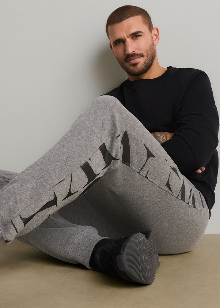 Er trägt ein schwarzes Oberteil, graue Jogginghose mit großem Schriftzug und schwarze Sneaker.