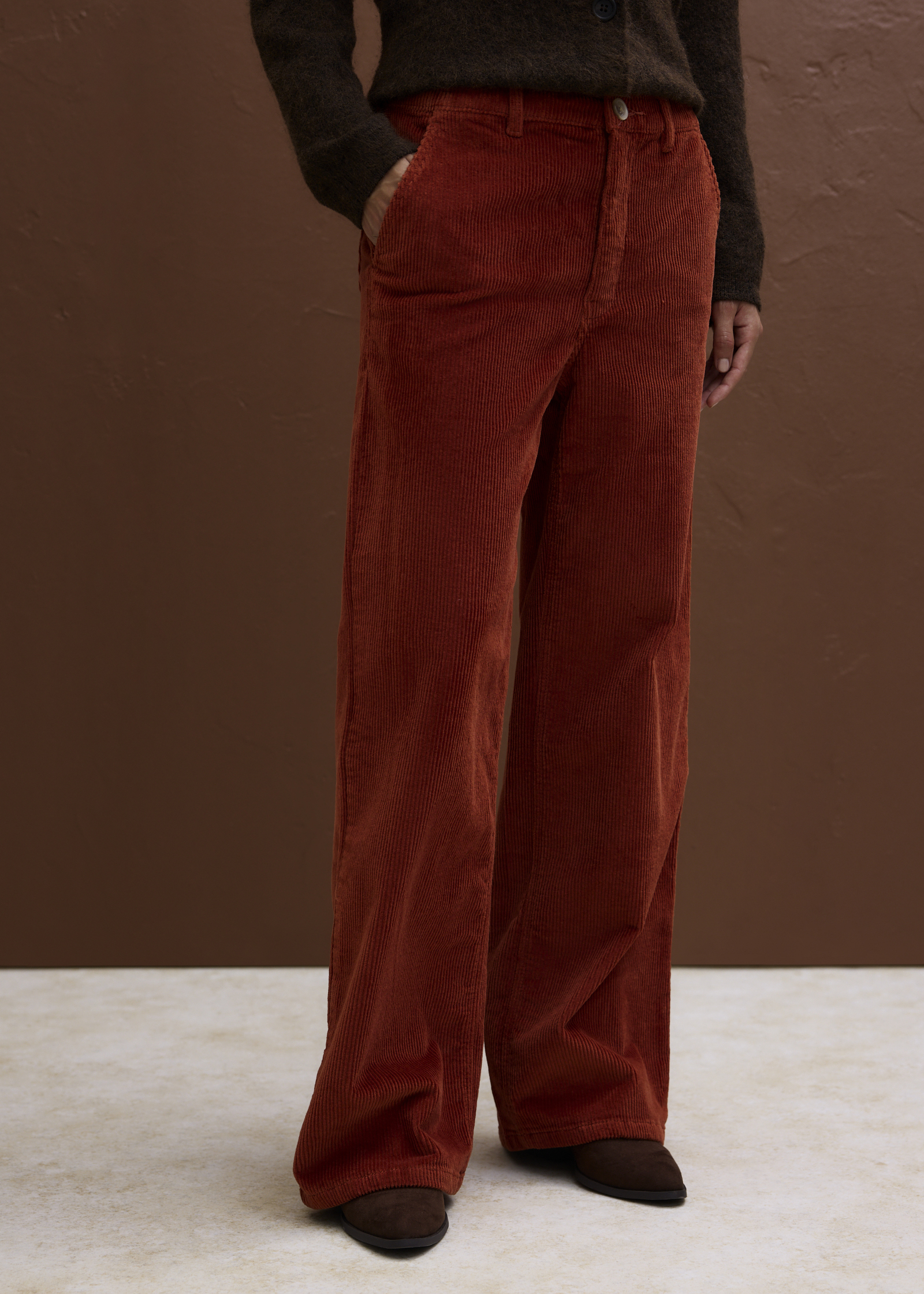 Sie trägt eine gemusterte Strickjacke über einem weißen Hemd und eine orange Cordhose.