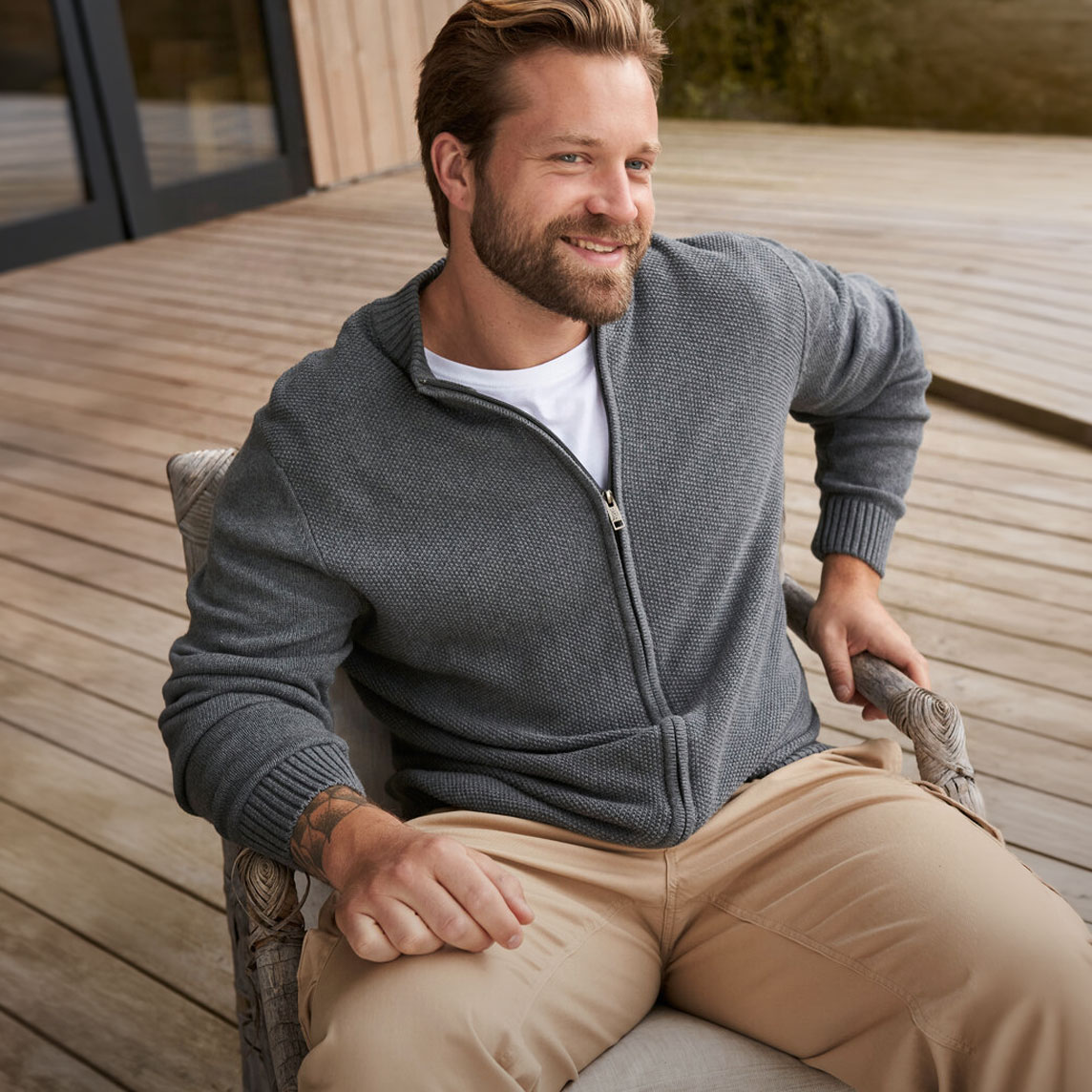 Er trägt eine graue Strickjacke über einem weißen T-Shirt und beige Hose.