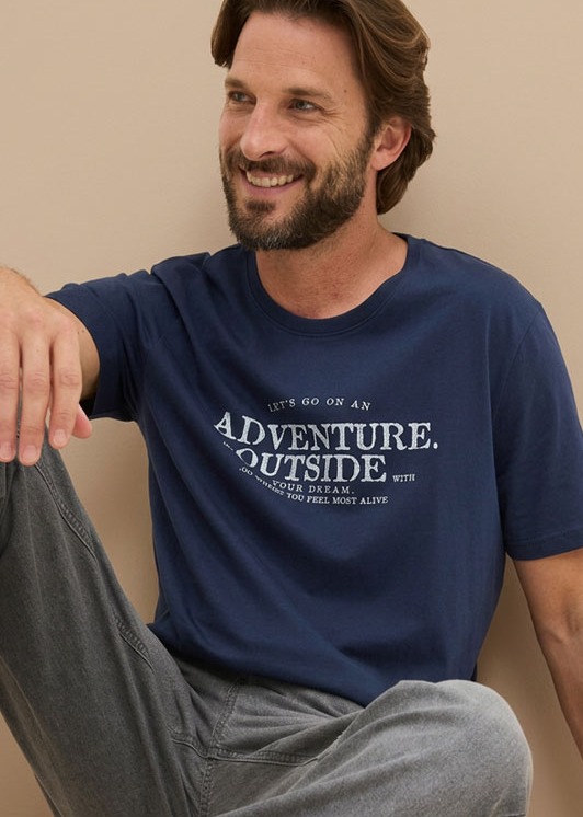Der Mann trägt ein dunkelblaues T-Shirt mit Aufdruck und eine graue Hose.
