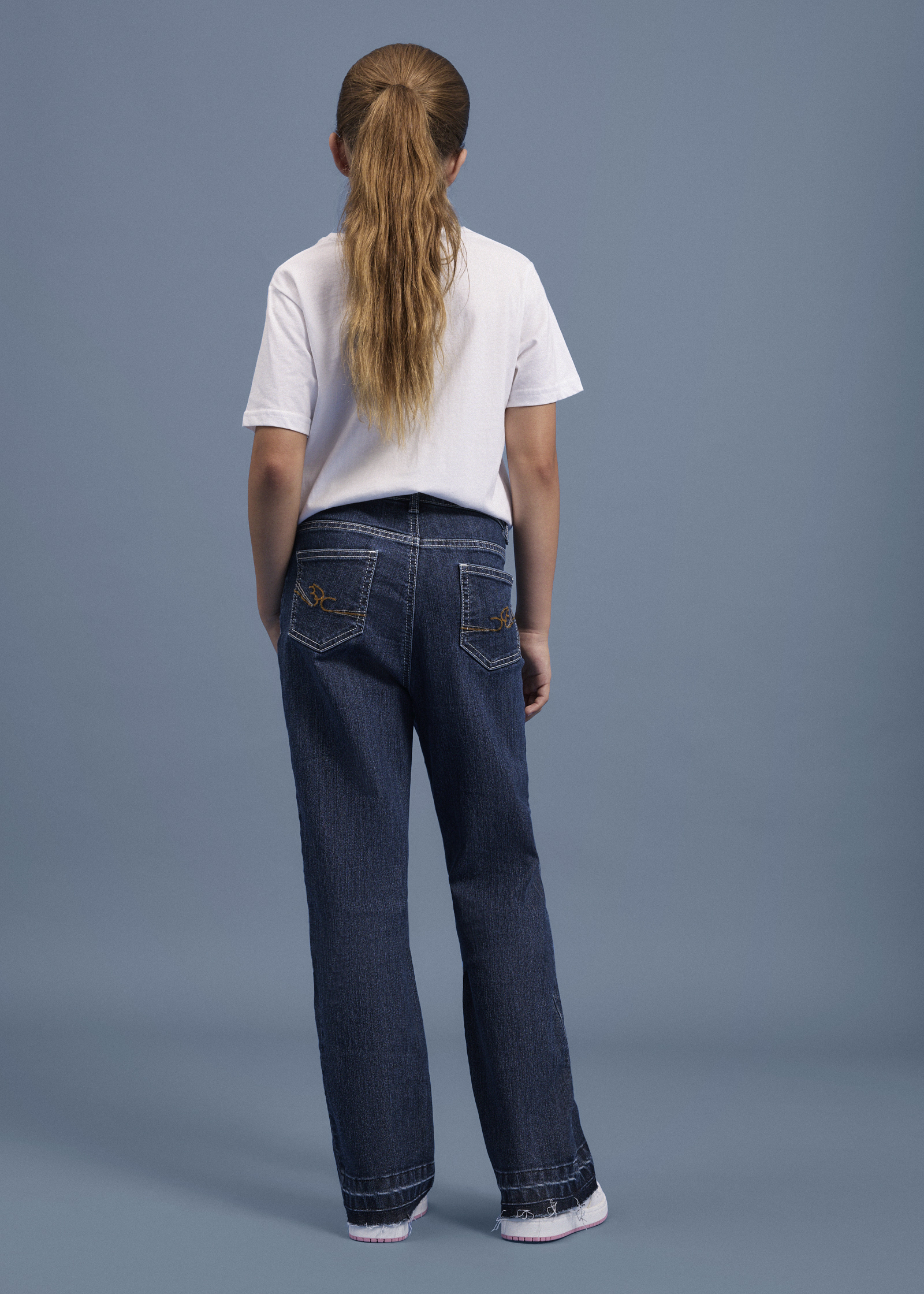 Ein Mädchen von hinten in einem weißen T-Shirt und blauen Jeans mit weißen Nähten.