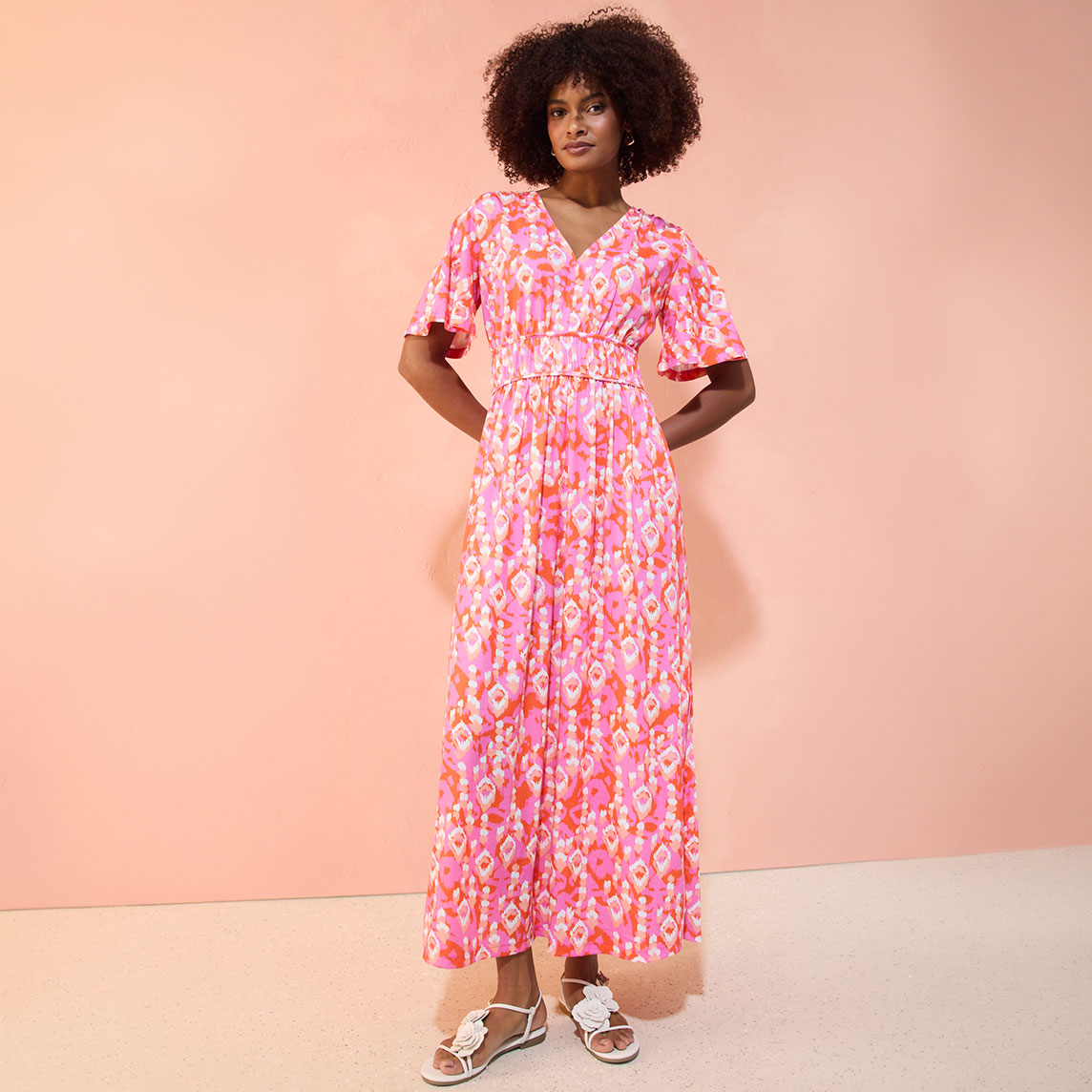 Eine Frau trägt ein langes, rosa gemustertes Kleid mit kurzen Ärmeln und weißen Sandalen.