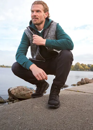 Er trägt eine grüne Kapuzenjacke, melierte Weste, dunkle Hose und feste Stiefel.