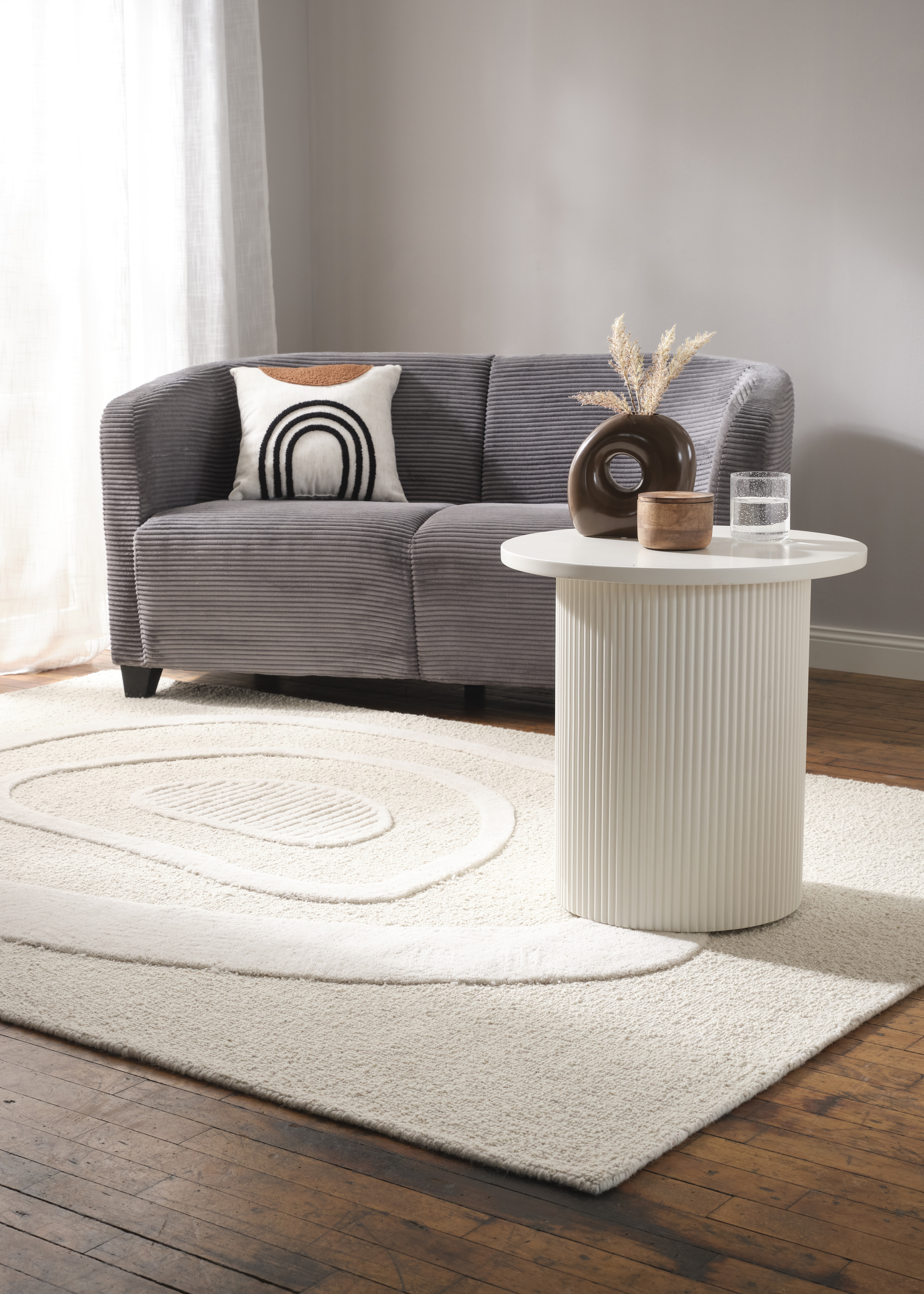 Gemütliches Wohnzimmer mit grauem Sessel, Rattanmöbeln und dekorativer Pflanze auf hellem Teppich.