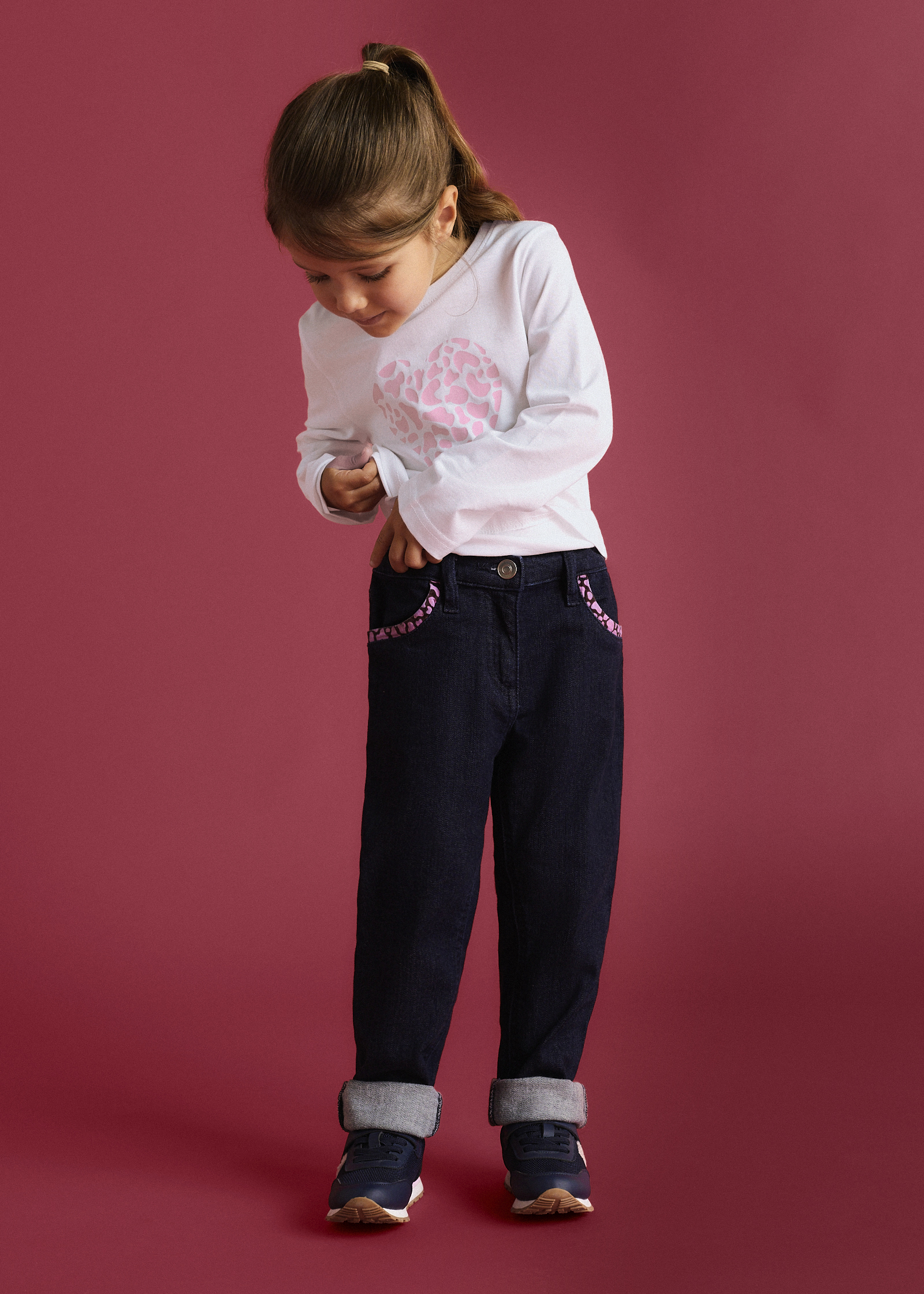 Weißes Herz-Shirt, dunkle Jeans mit gemusterten Taschen und umgeschlagenem Saum, dazu Sneakers.
