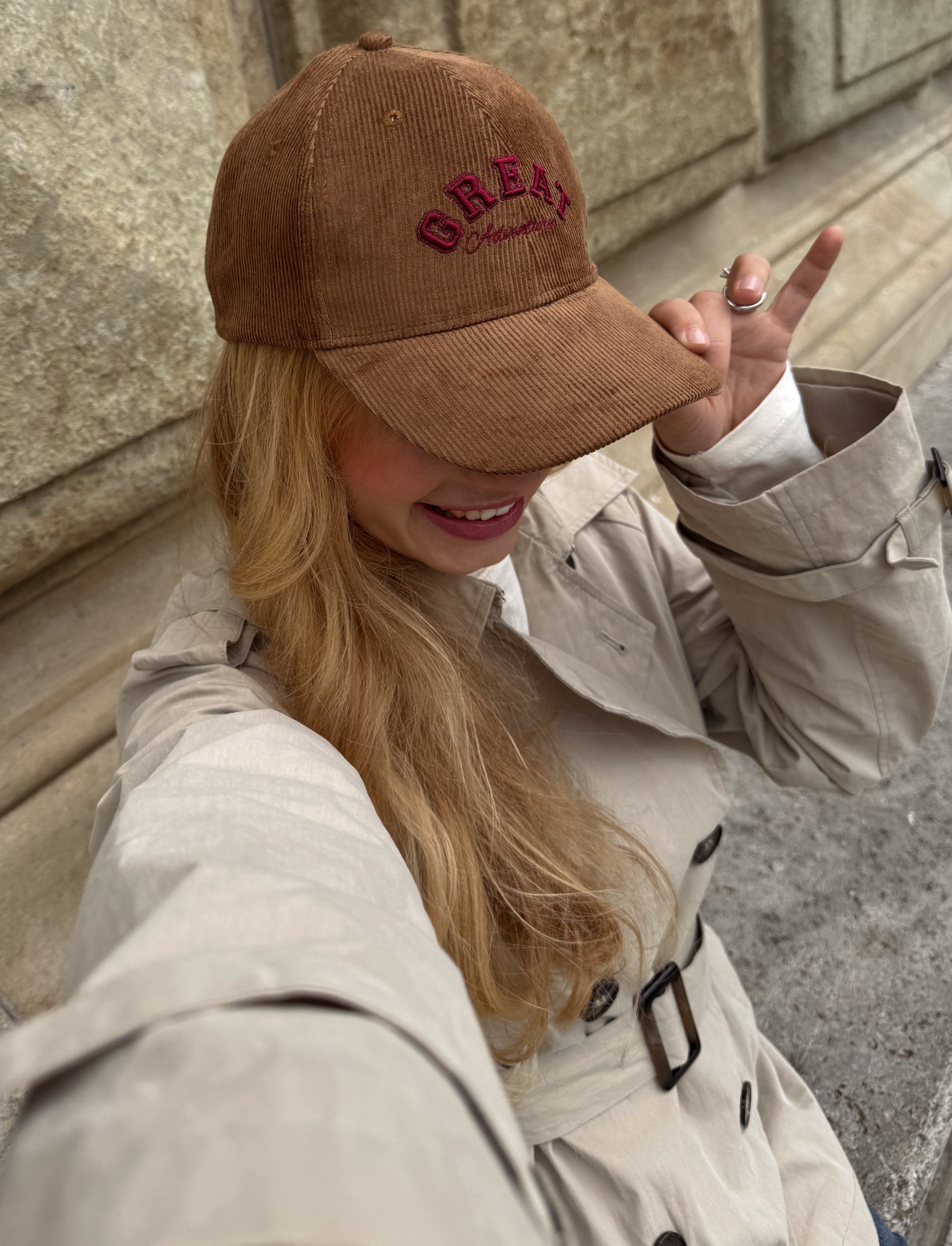 Beiger Trenchcoat und braune Baseballkappe mit roter Aufschrift.