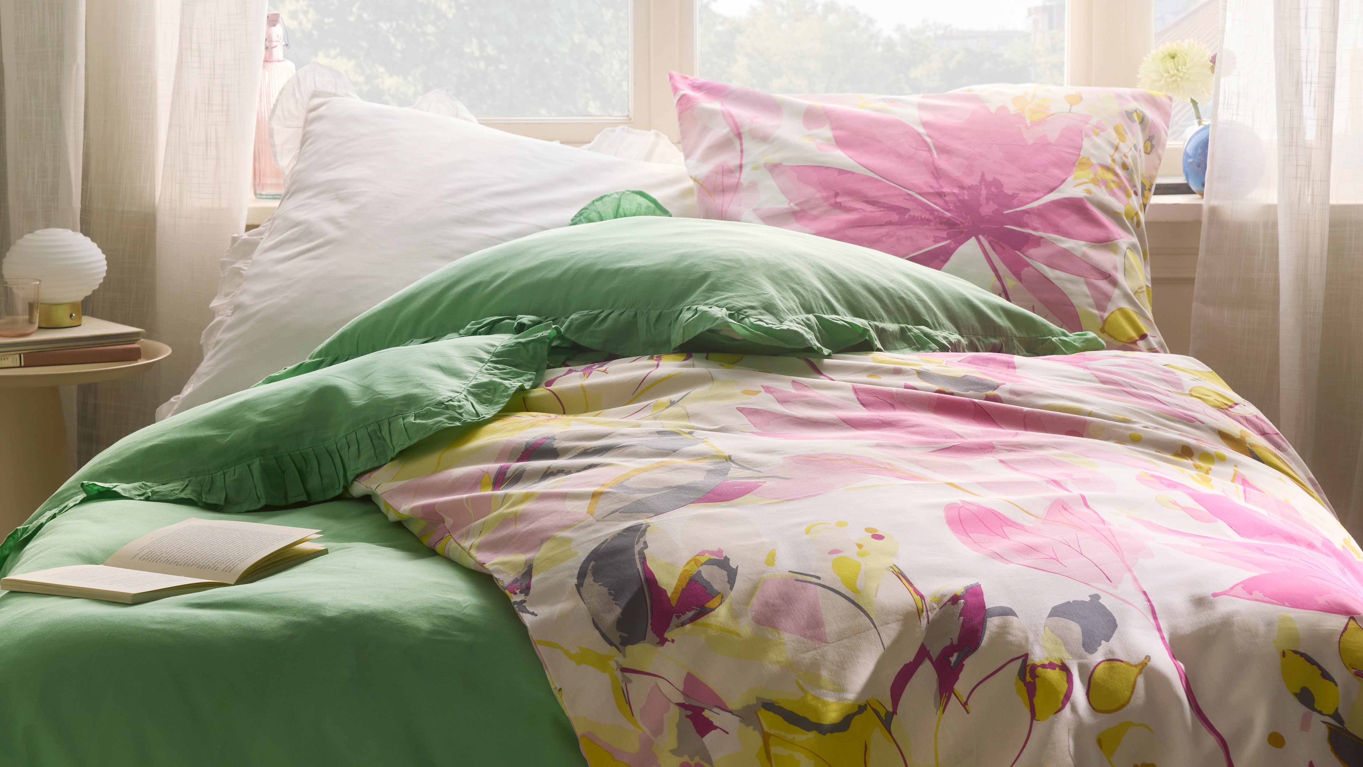 Farbenfrohe Bettwäsche mit Blumenmuster, kombiniert mit grünem Kissen. Zarte Vorhänge rahmen das Bett ein.