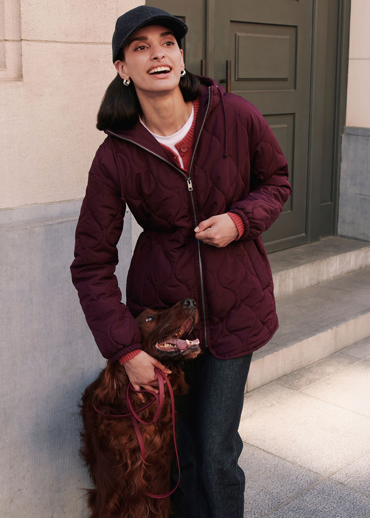 Dunkle Kappe, gesteppte bordeauxrote Jacke, roter Cardigan und dunkle Jeans.