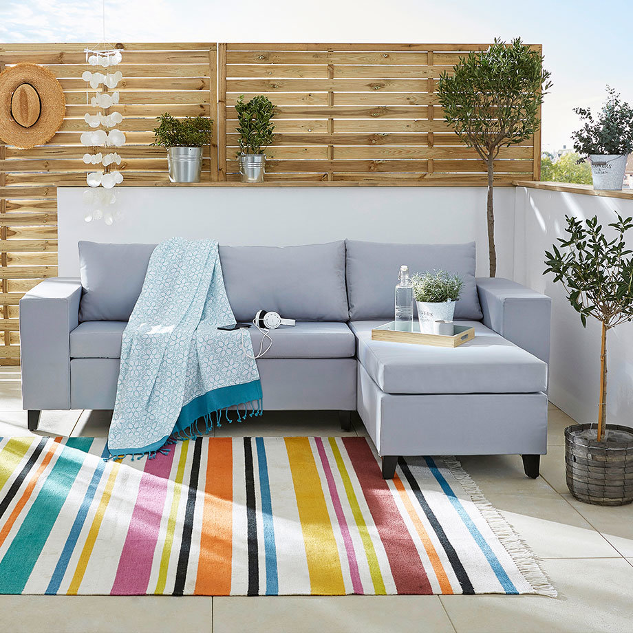 Graues Sofa mit hellblauer Decke auf buntem Teppich. Gemütliche Outdoor-Sitzecke für Sommertage.