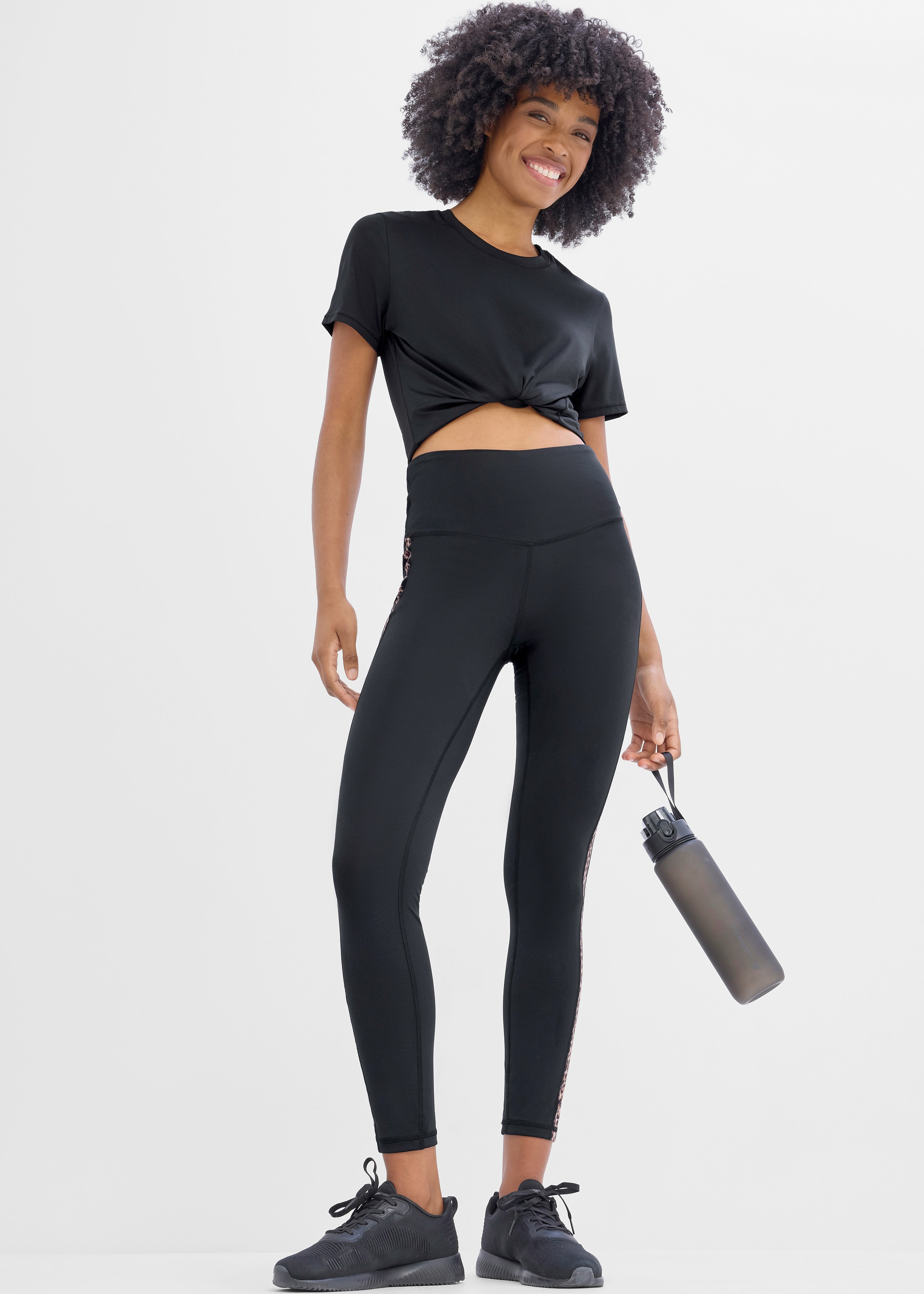 Modisches Sportoutfit: Schwarzes, geknotetes Oberteil, Leggings und bequeme schwarze Turnschuhe.