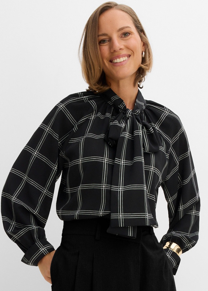 Elegante schwarze Bluse mit Karomuster und Schleife, schwarze Hose, goldener Armreif.