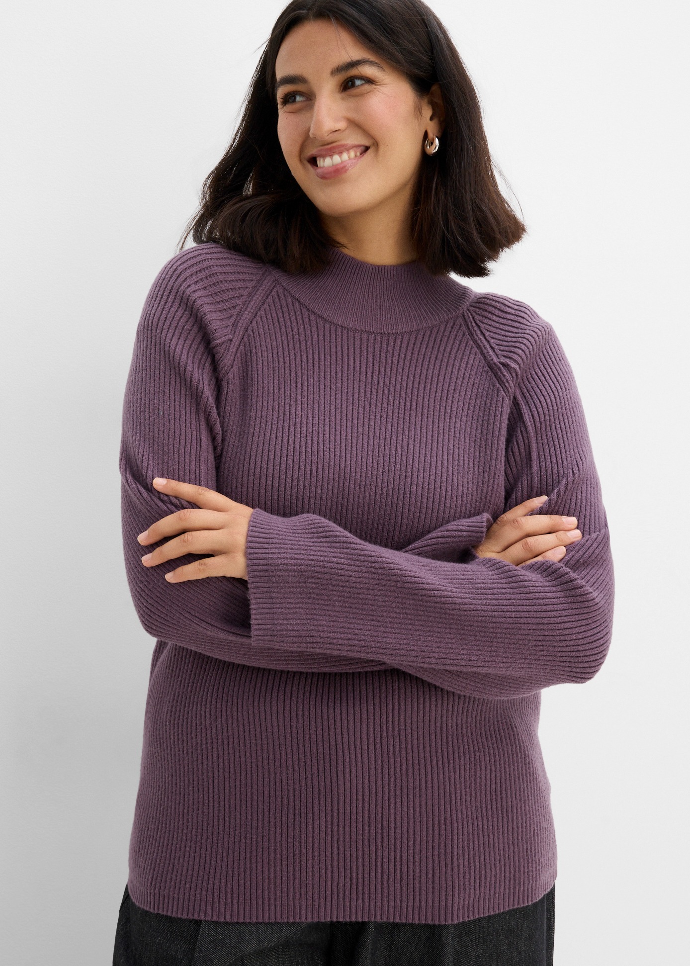Ein gerippter, lila Strickpullover mit hohem Kragen und dunkle Hose.