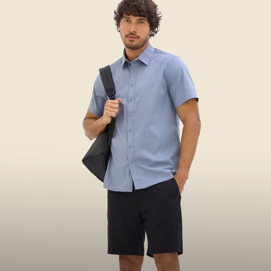 Der Mann trägt ein hellblaues Kurzarmhemd, schwarze Shorts und eine Umhängetasche.