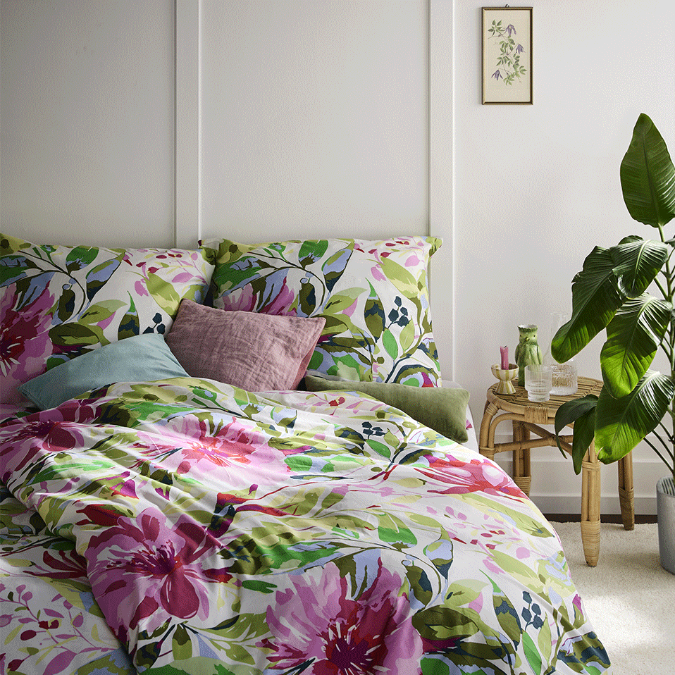 Farbenfrohe Blumenmuster zieren die Bettwäsche, ergänzt durch ein lilafarbenes Kissen und weiße Wände.