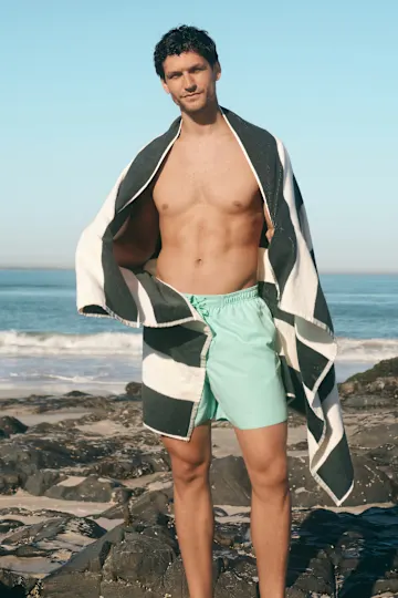 Mann trägt mintgrüne Badeshorts und stylisches gestreiftes Strandtuch über den Schultern.