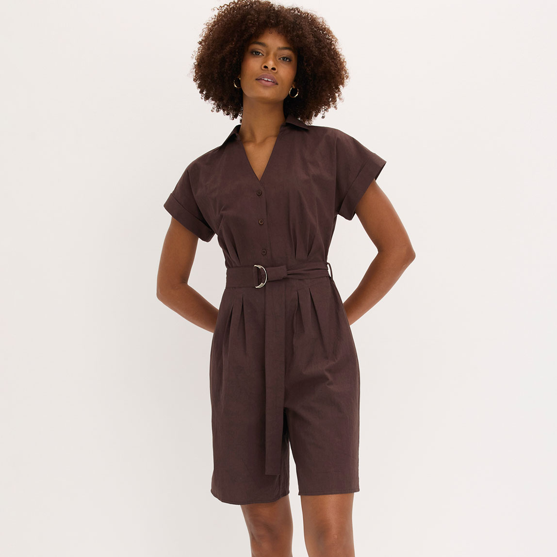 Die Frau trägt einen eleganten braunen Jumpsuit mit Gürtel und kurzen Ärmeln.