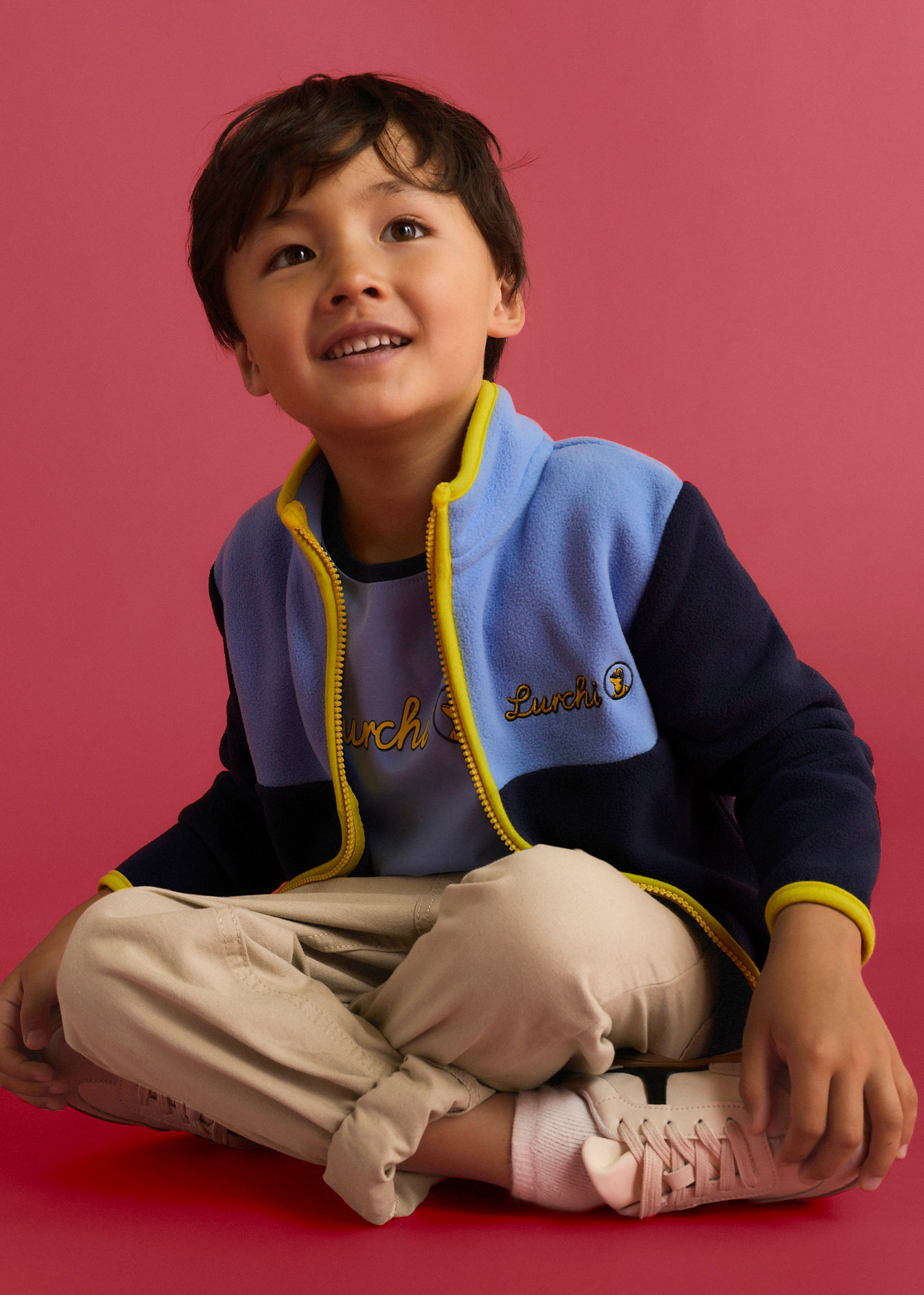 Der Junge trägt eine bunte Fleecejacke, ein hellblaues Shirt, beige Hose und helle Turnschuhe.