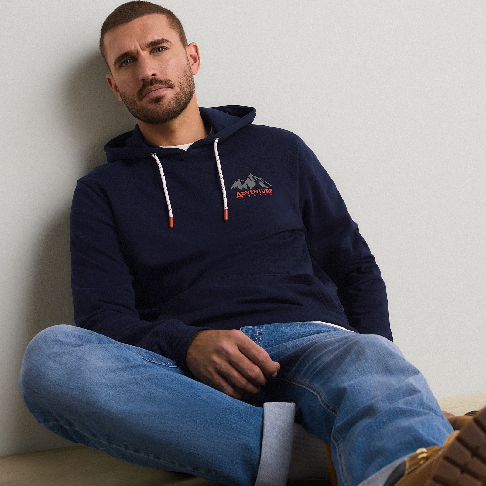 Er trägt einen dunkelblauen Hoodie mit Bergmotiv und dazu eine lässige blaue Jeans.