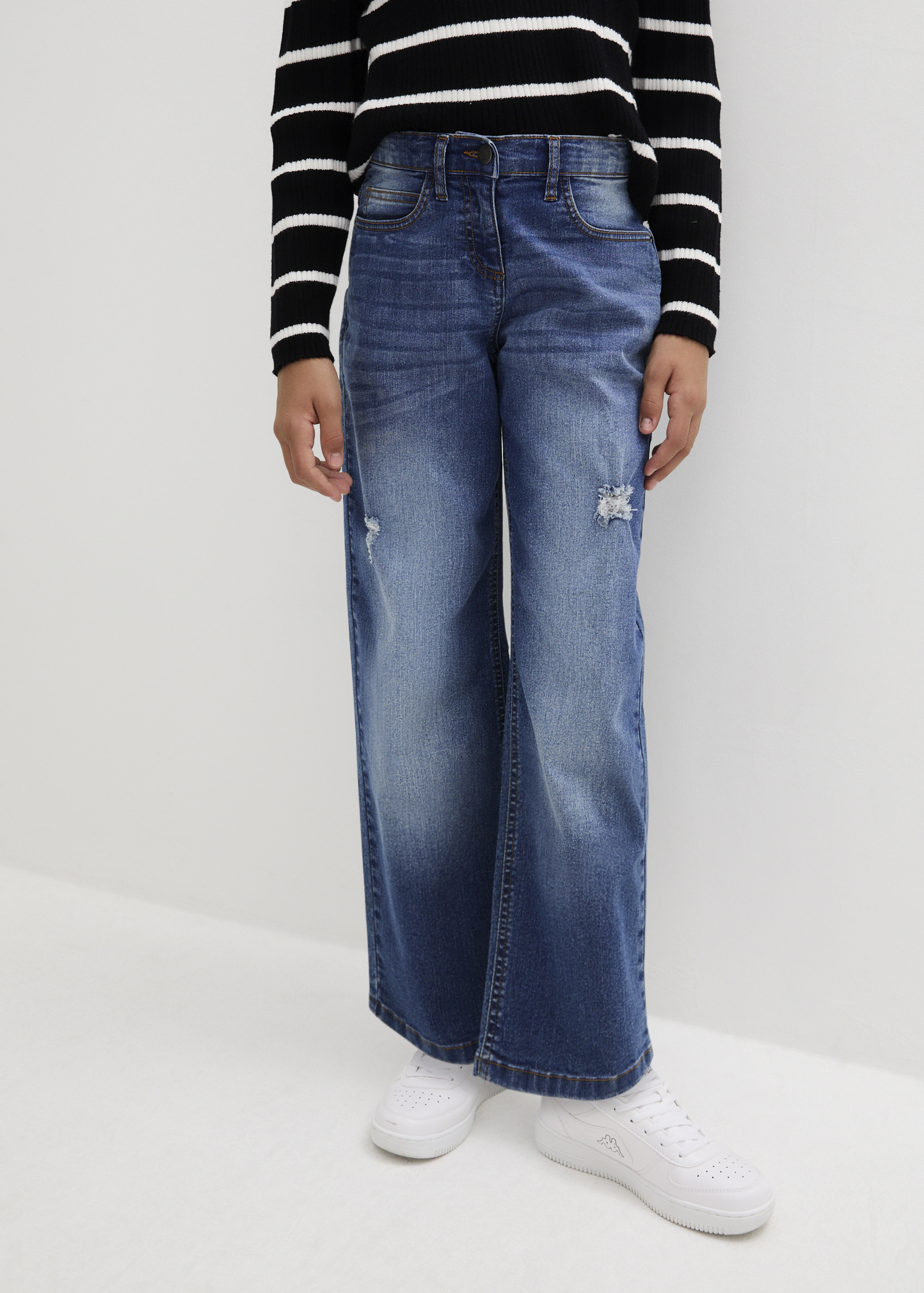 Das Kind trägt gestreiftes Langarmshirt, blaue Schlagjeans und weiße Sneaker.