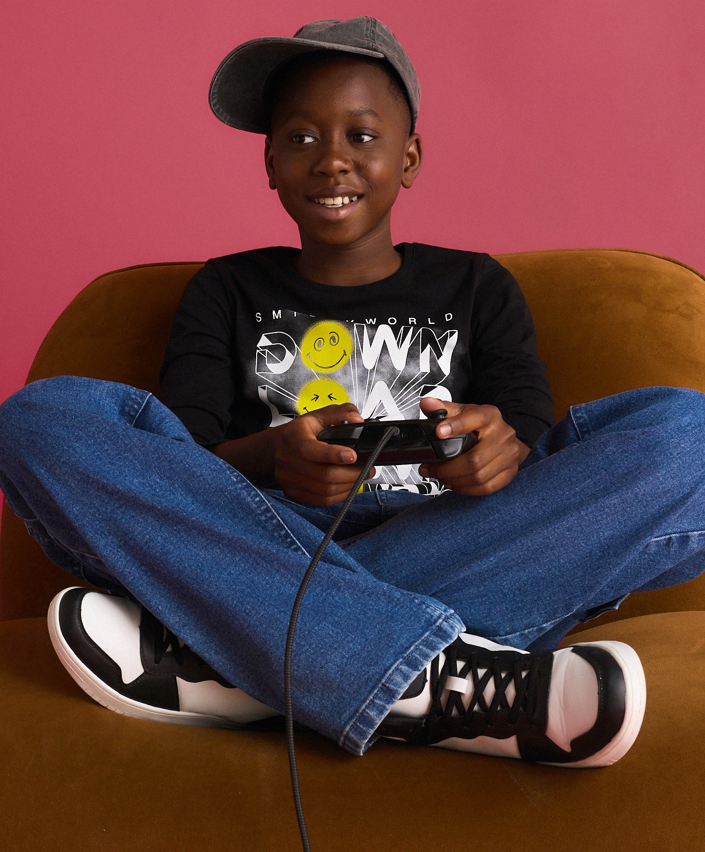 Kind trägt graue Kappe, schwarzes Shirt mit Smileys, weite Jeans und trendige Sneaker.