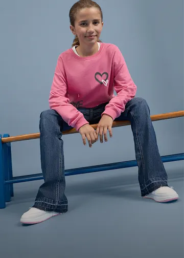 Sie trägt ein rosa Sweatshirt mit Herzen, blaue Jeans und weiße Turnschuhe.
