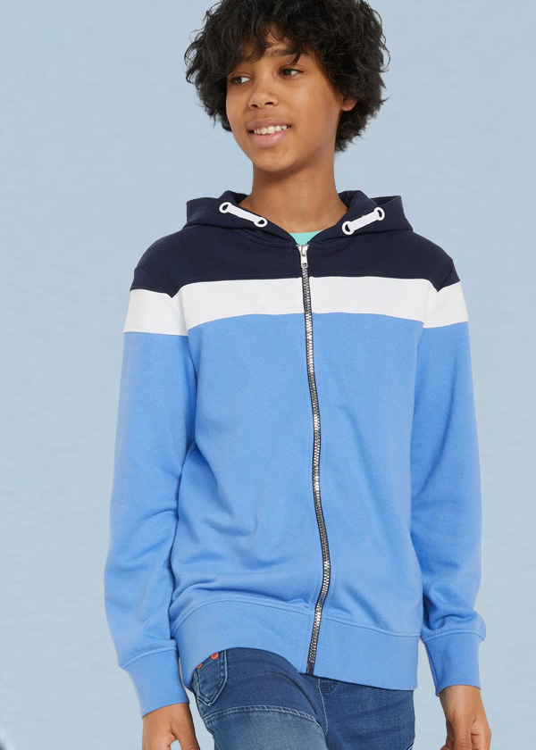Der Junge trägt eine gestreifte Kapuzenjacke in Blautönen, Weiß und Navy mit Jeans.