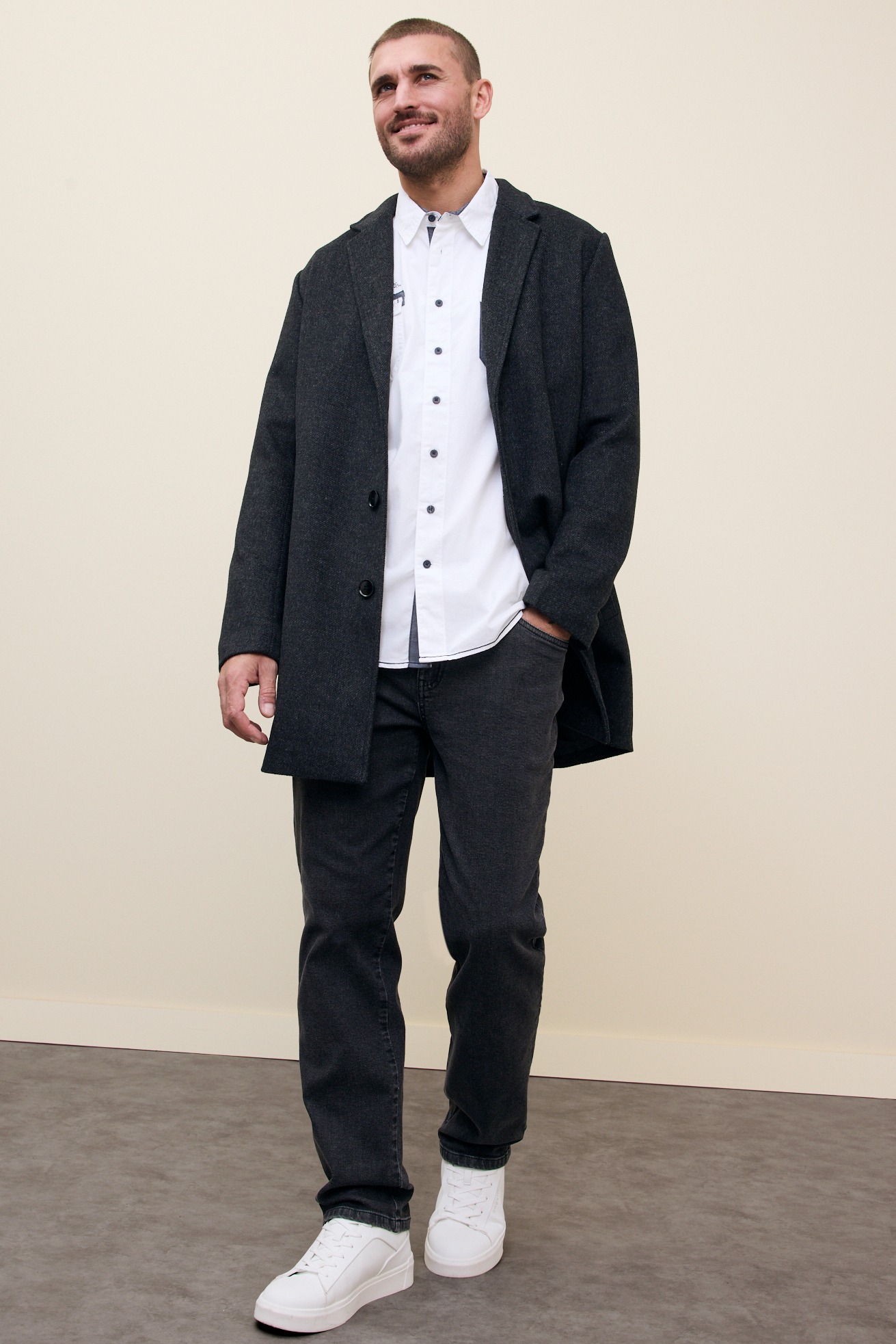 Dunkler Mantel über weißem Hemd, passende Jeans und weiße Sneaker.
