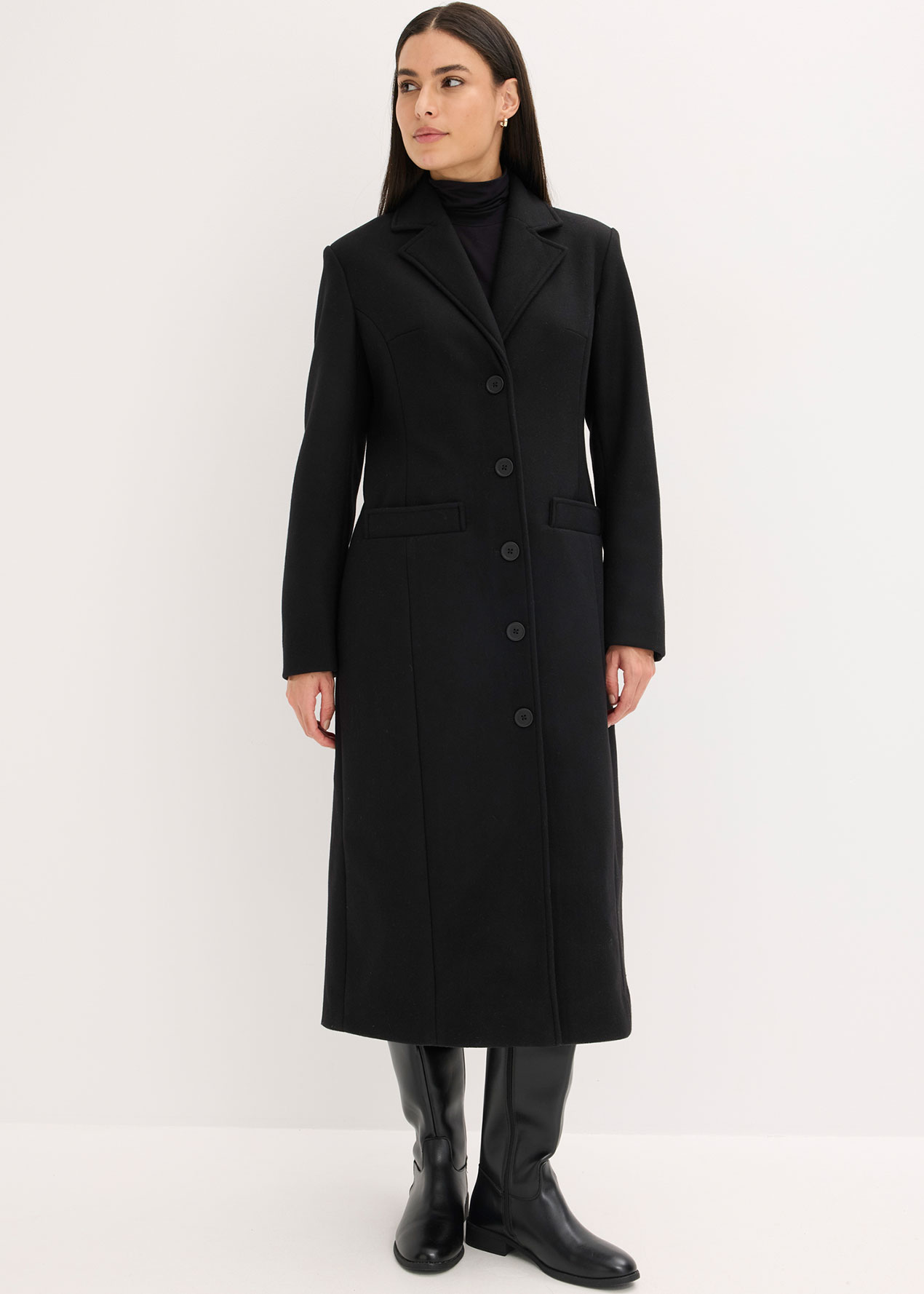 Sie trägt einen langen schwarzen Mantel, einen Rolli und passende schwarze Stiefel.