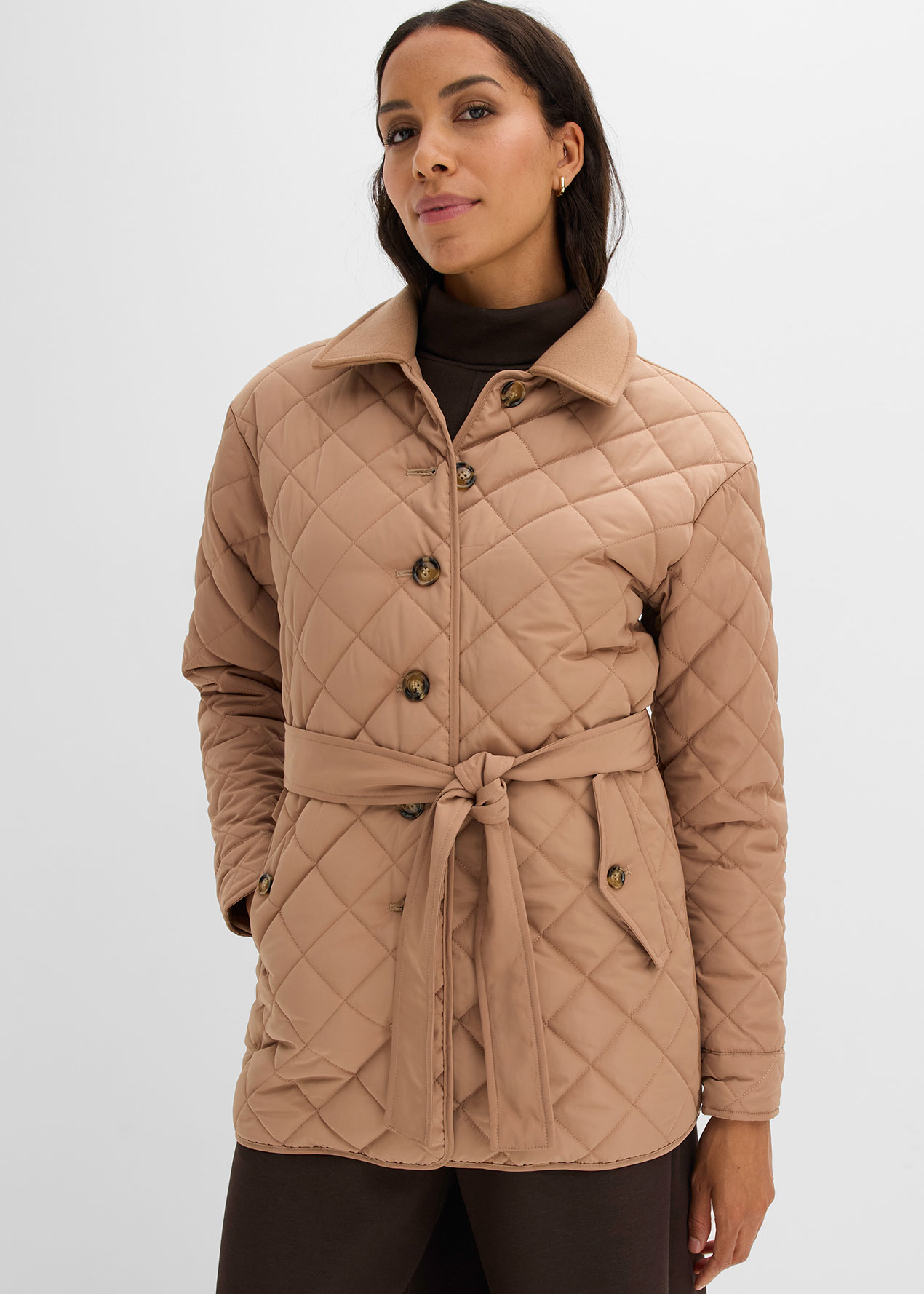 Die stilvolle beigefarbene Steppjacke mit Gürtel wird über einem Rollkragenpullover getragen.