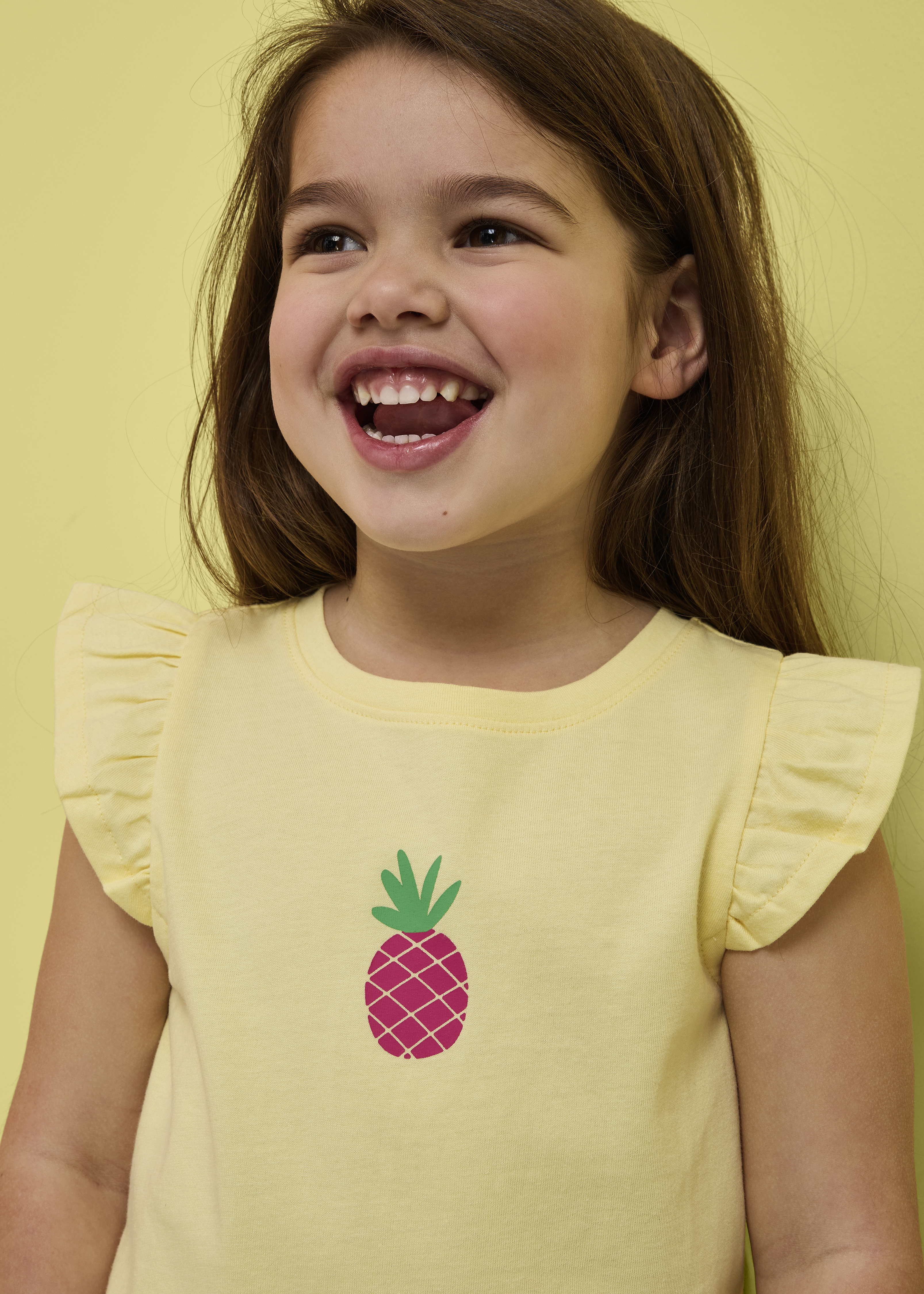 Mädchen trägt ein gelbes T-Shirt mit Ananas-Motiv und Rüschenärmeln.