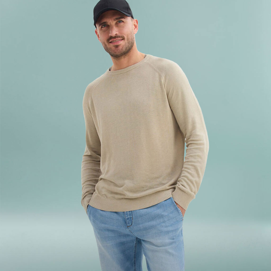 Mann trägt beigen Pullover, blaue Jeans und schwarze Baseballkappe.