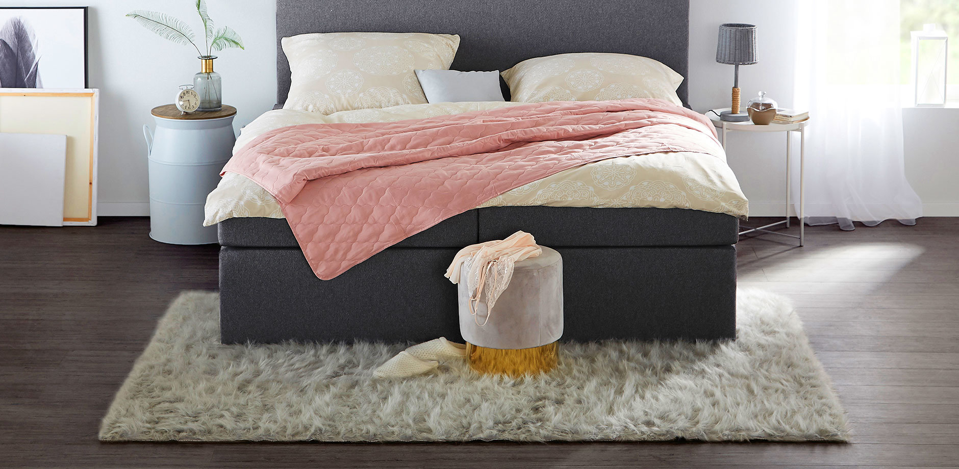 Gemütliches Schlafzimmer mit grauem Bett, rosa Decke und flauschigem Teppich. Helle Bettwäsche sorgt für Kontrast.