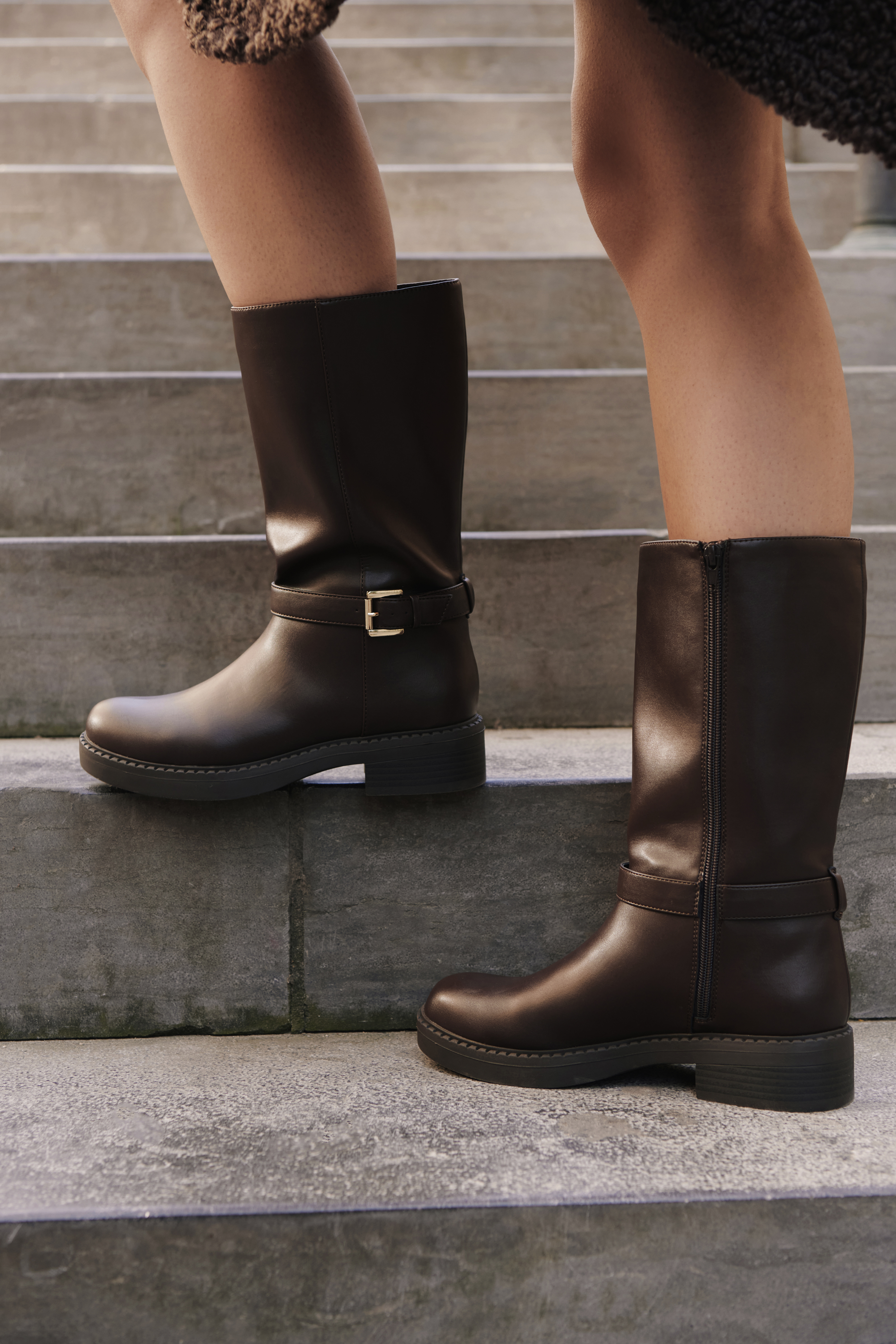 Dunkelbraune Stiefel mit goldener Schnalle und Profilsohle auf steinernen Stufen.