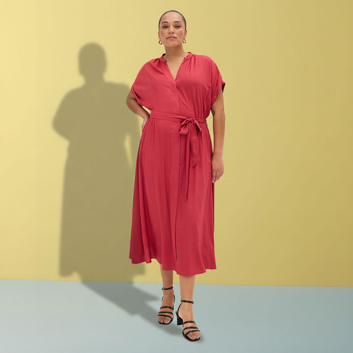 Elegantes rotes Wickelkleid mit Gürtel und Sandalen vor gelber Wand.