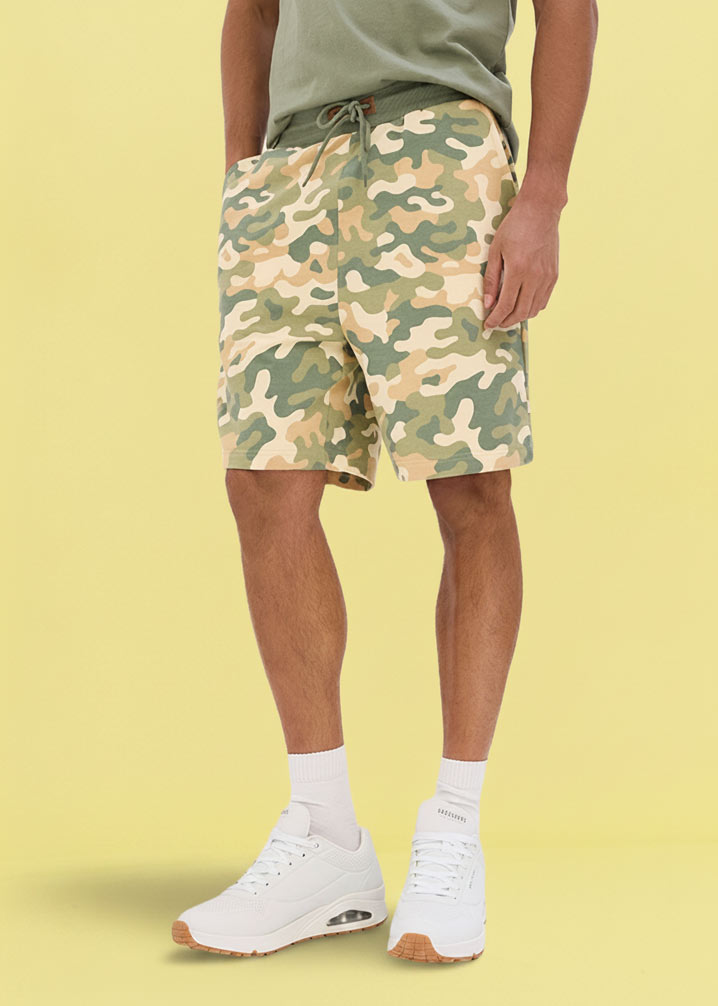 Lässige Camouflage-Sweat-Shorts in Grün-Braun-Tönen kombiniert mit hellem T-Shirt und weißen Sneakern.
