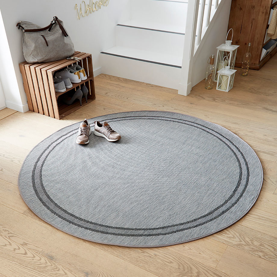 Grauer runder Teppich und weiße Sneakers auf hellem Holzboden. Schicke Handtasche auf Holzregal.