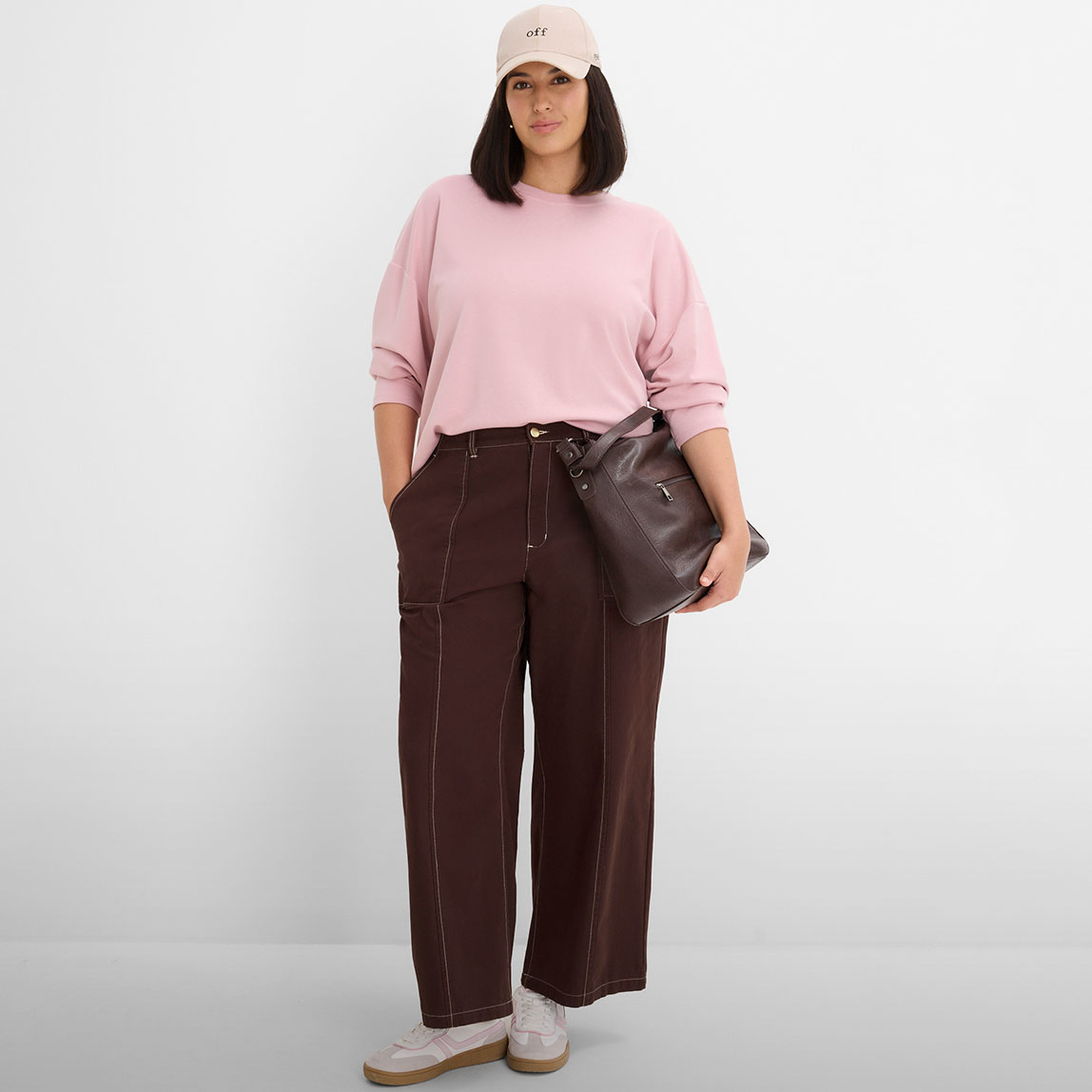 Rosa Sweatshirt, braune Weithose mit Nähten, beiges Cap und dunkle Tasche.